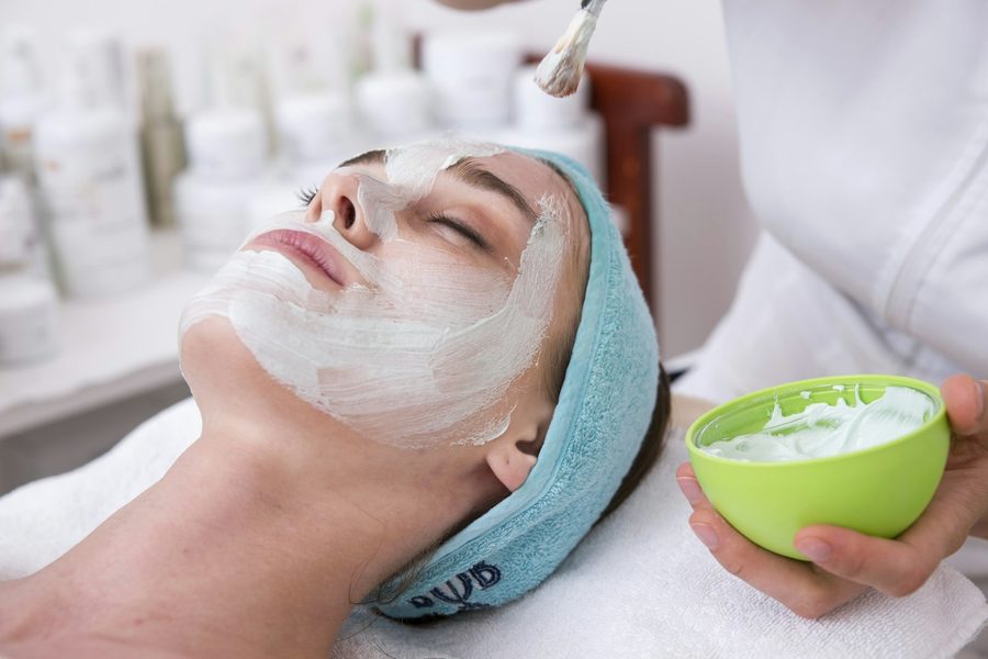 Clienta con mascarilla facial blanca mientras la esteticista sostiene el bol del producto