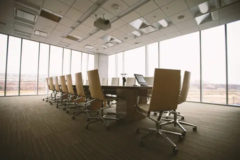 Sala de reuniones vacía con una mesa larga, sillas de oficina y ventanales del suelo al techo