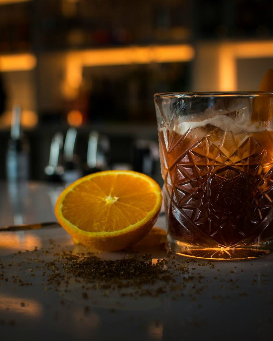 Vaso tallado con un cóctel ámbar y media naranja sobre la barra en penumbra.