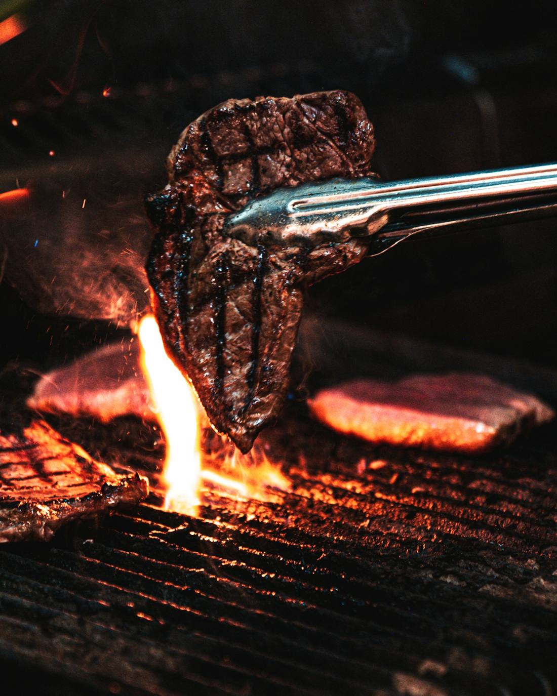 Unas pinzas levantan un chuletón marcado sobre la parrilla, con una llama debajo.