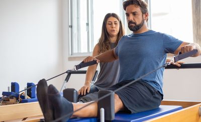Persona trabajando en una máquina reformer de pilates mientras la instructora corrige la postura